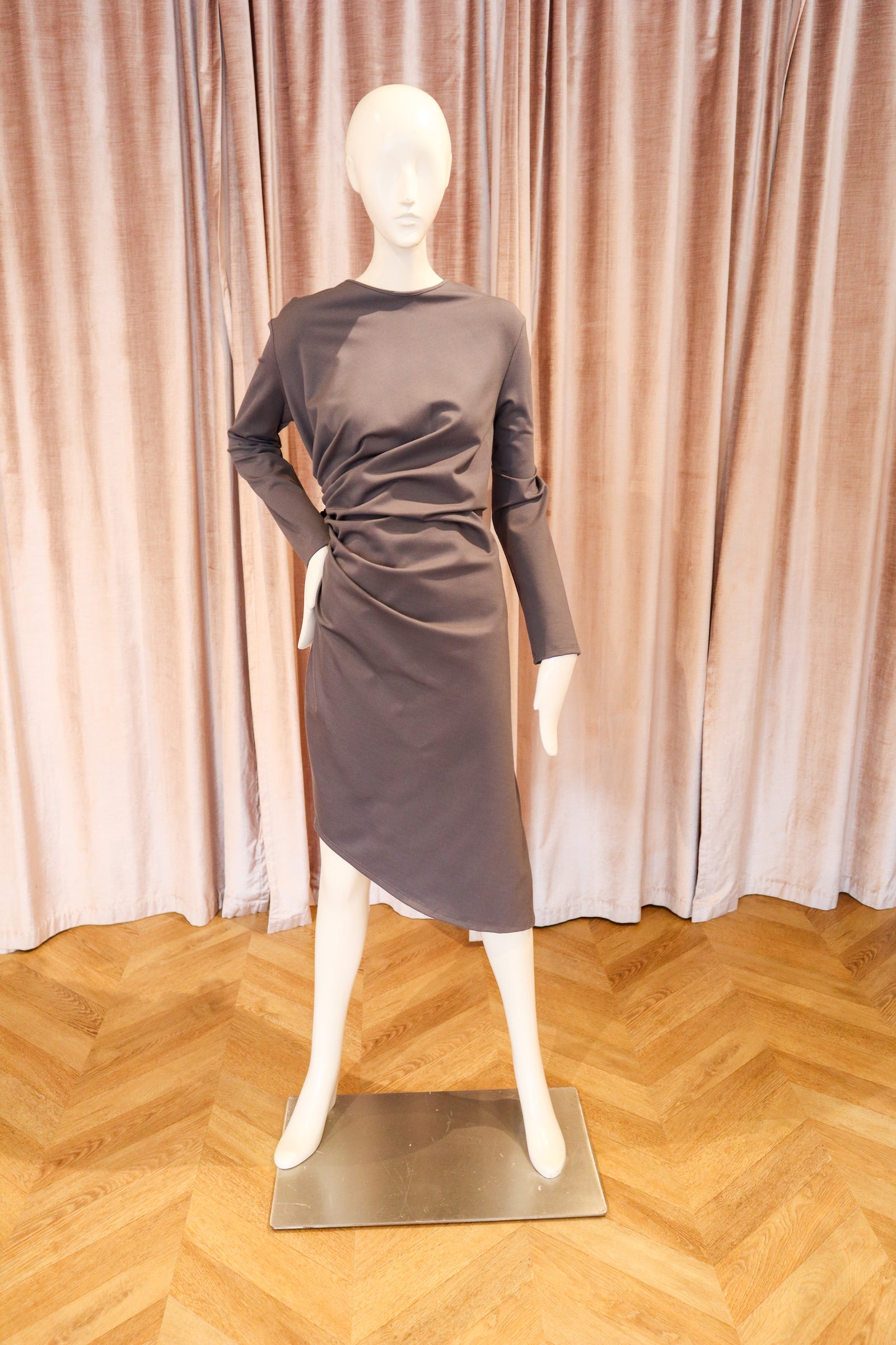 A dark graylong-sleeve dress displayed on a white mannequin, set against a backdrop of pinkish-beige velvet curtains and a light wood herringbone floor.