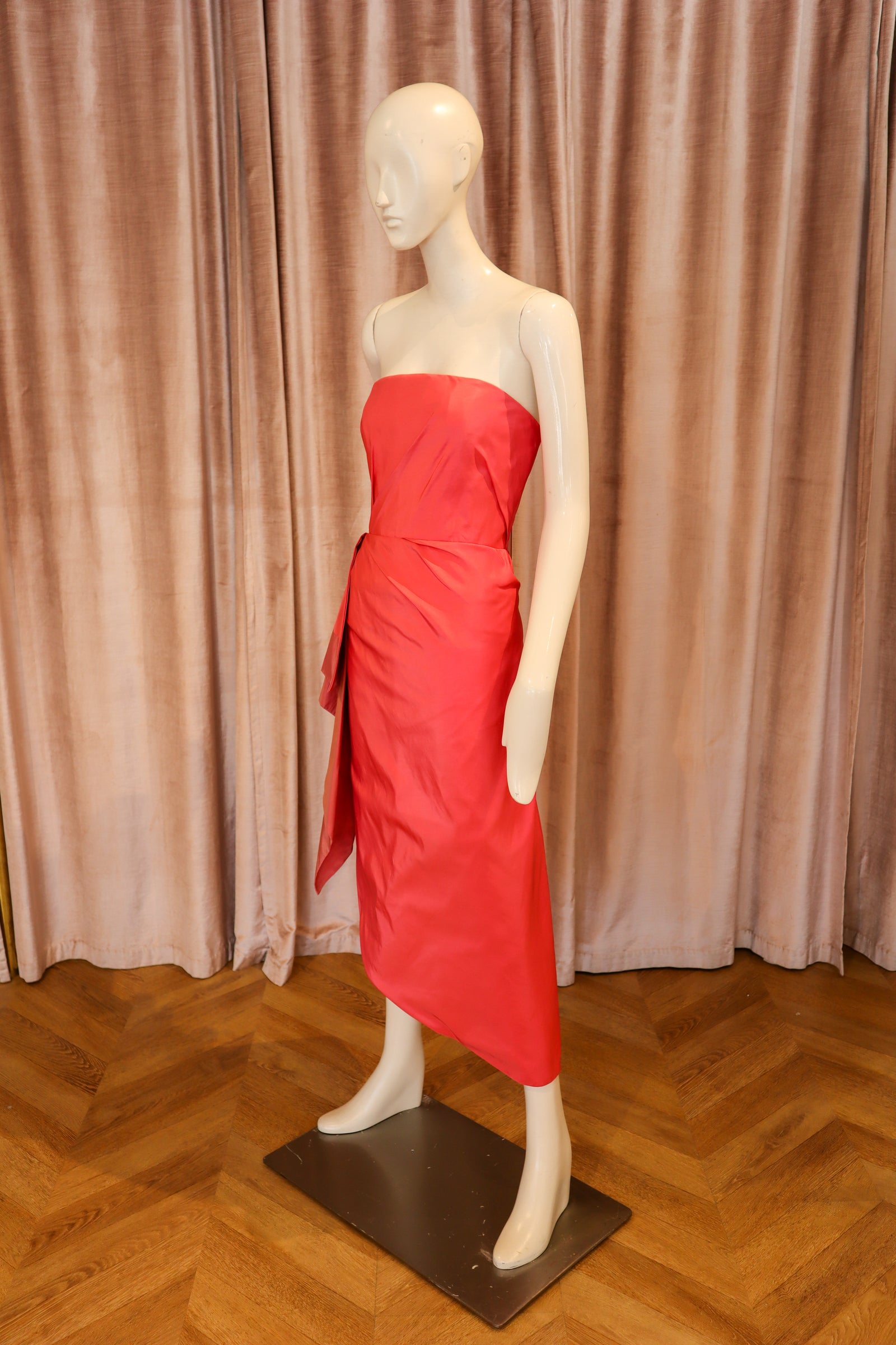 A bright red strapless midi dress with a draped, layered asymmetrical hem, displayed on a female mannequin against a pinkish-brown velvet curtain and wooden parquet floor.