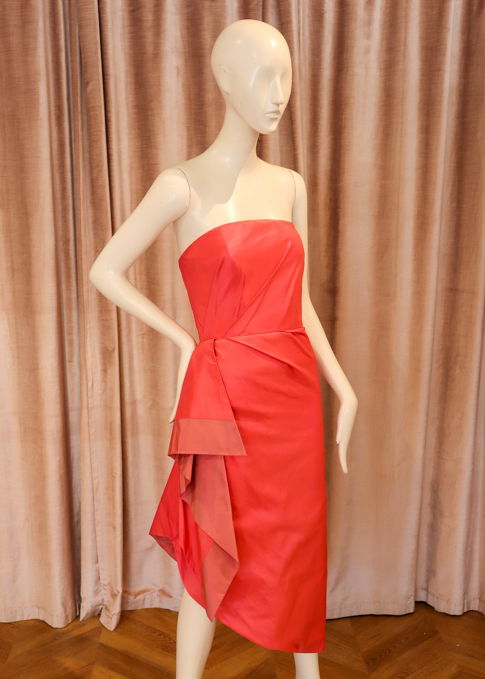A bright red strapless midi dress with a draped, layered asymmetrical hem, displayed on a female mannequin against a pinkish-brown velvet curtain and wooden parquet floor.