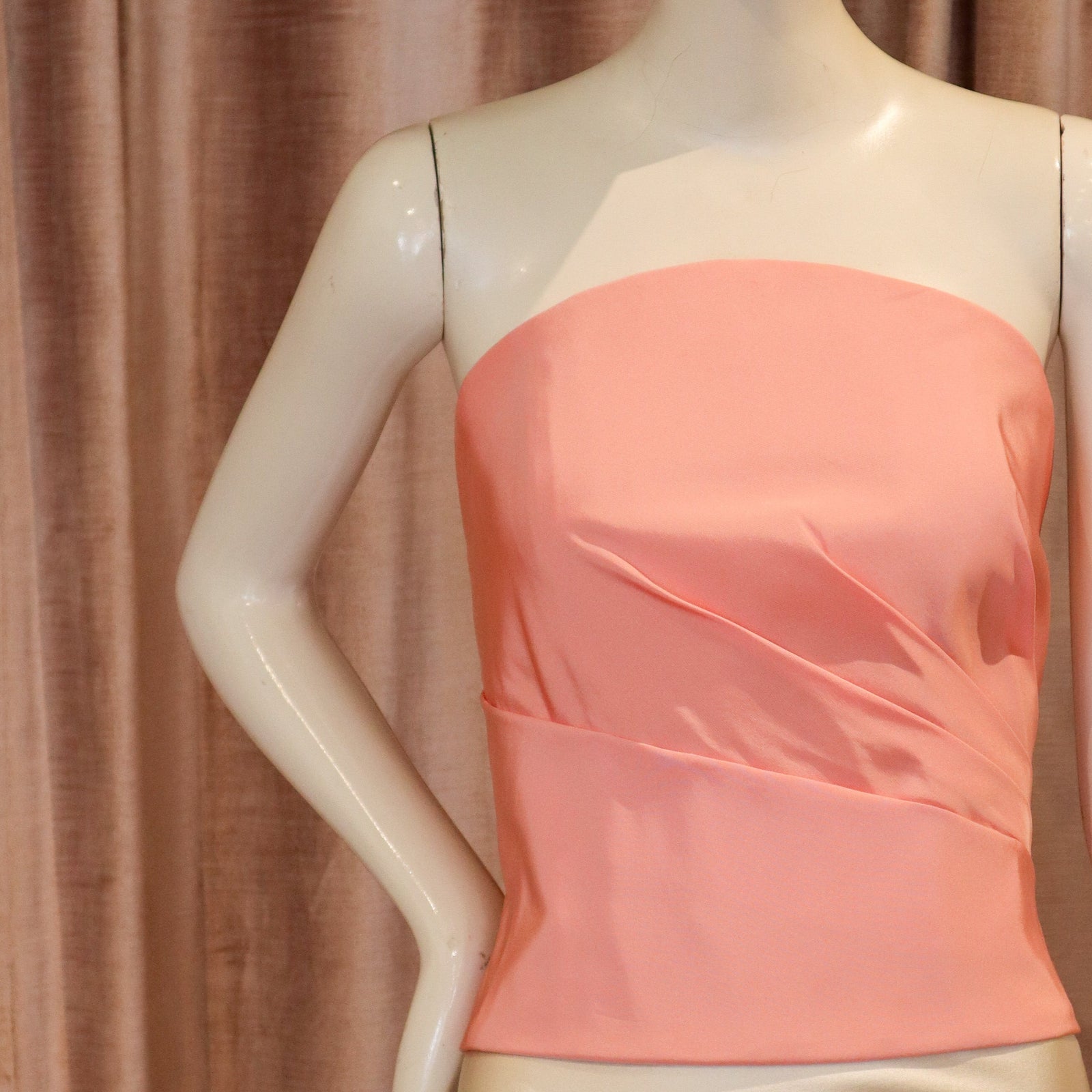A peach-pink strapless satin top with draped side details, displayed on a cream-colored mannequin against a brown curtain backdrop.