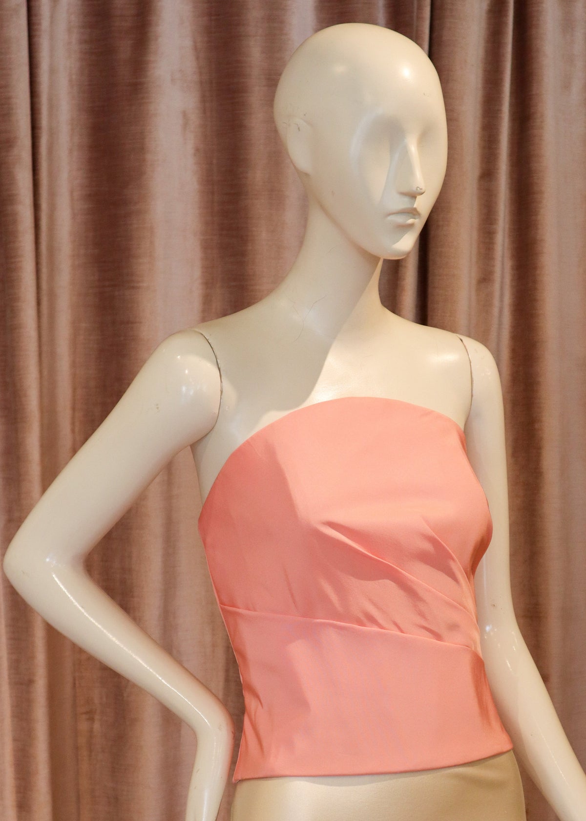 A peach-colored strapless bustier top displayed on a cream mannequin, paired with a beige satin skirt, set against a brown curtain backdrop.