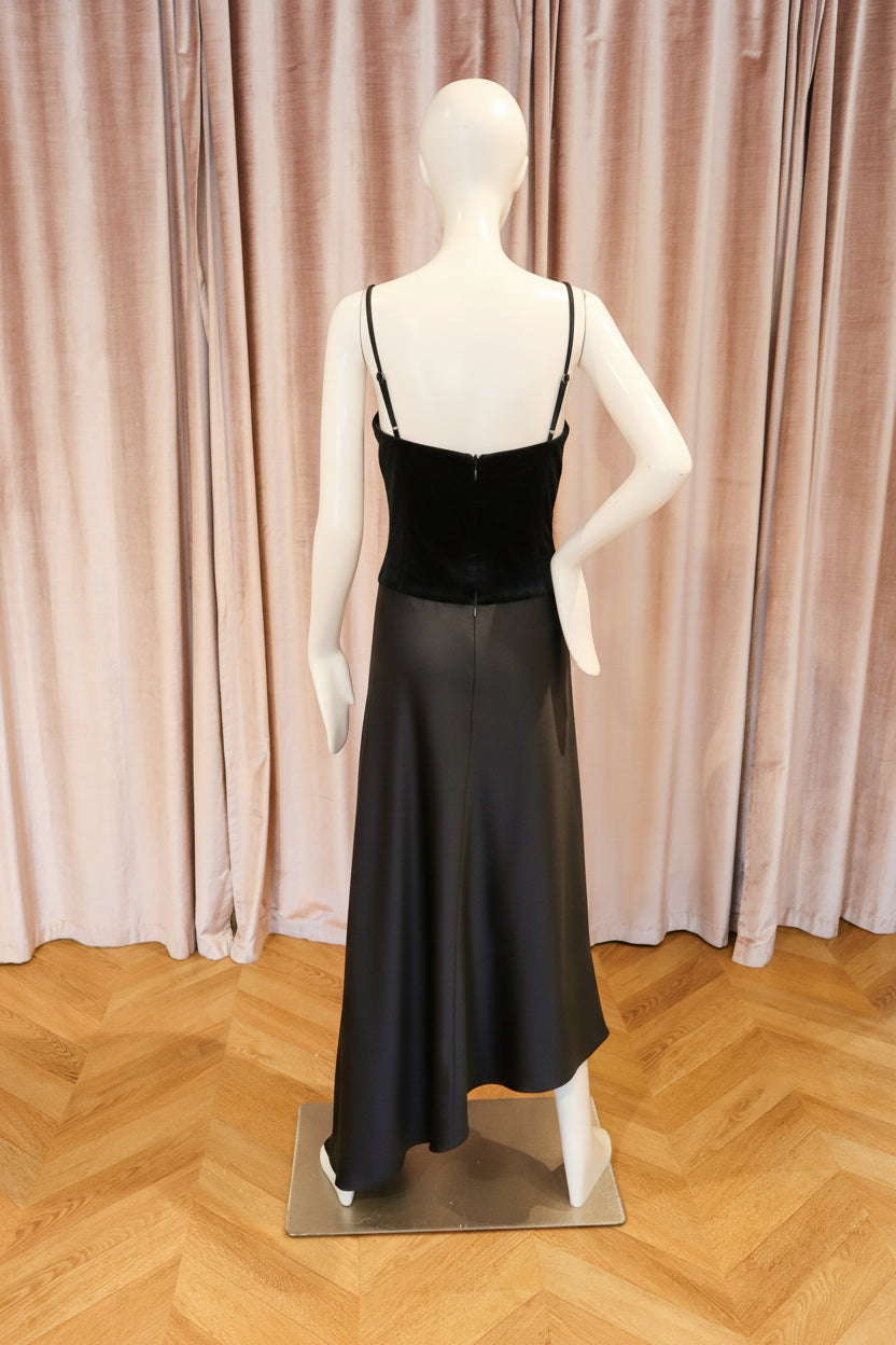 A strapless satin midi dress in a rich black color, displayed on a white mannequin from the back. The dress features a structured corset-style bodice and a flowing skirt, set against a background of pinkish-beige curtains and a light wood herringbone floor.