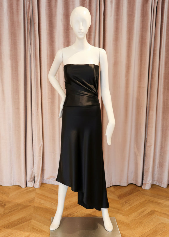 A strapless satin midi dress in a rich black color, displayed on a white mannequin standing on a wooden parquet floor in front of a pink velvet curtain.