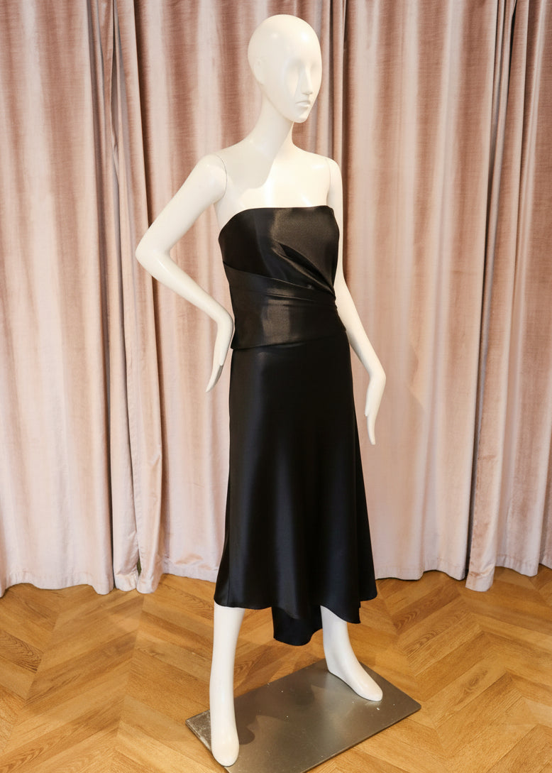 A strapless satin midi dress in a rich black color, displayed on a white mannequin standing on a metal base against a backdrop of pinkish-beige velvet curtains and a wooden parquet floor.