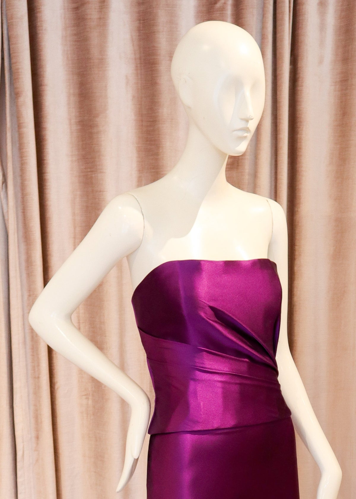 A strapless satin midi dress in a rich purple color, displayed on a white mannequin standing on a metal base against a backdrop of pinkish-beige velvet curtains and a wooden parquet floor.