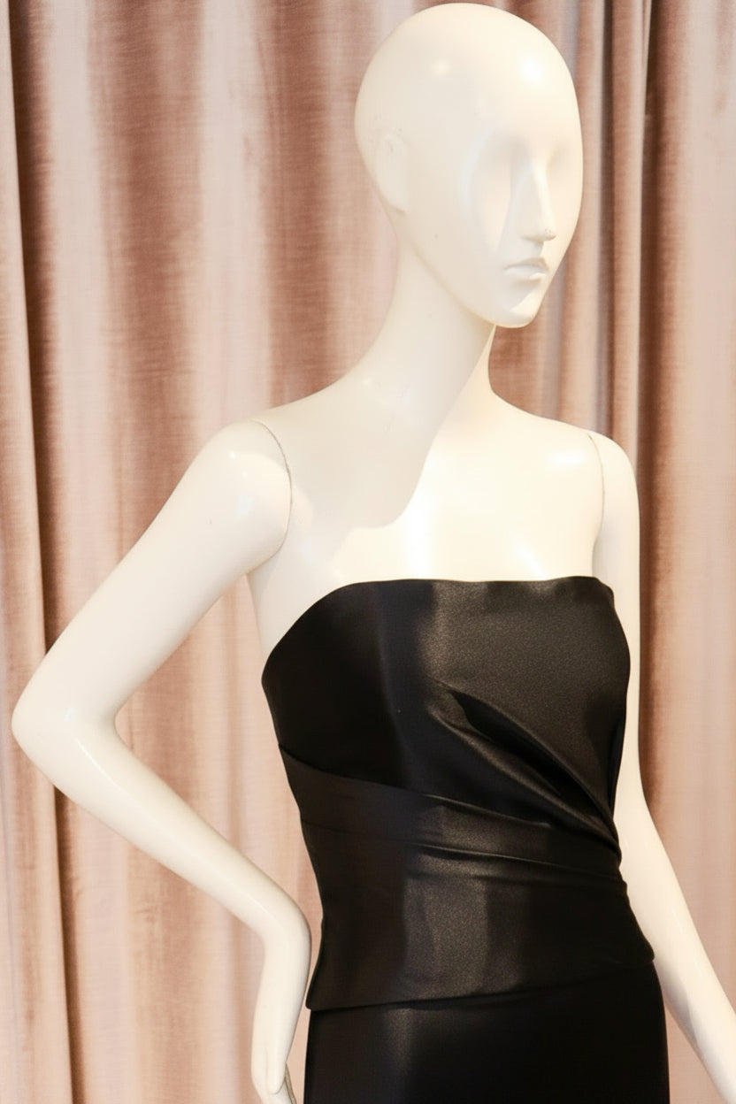 A strapless satin midi dress in a rich purple color, displayed on a white mannequin standing on a metal base against a backdrop of pinkish-beige velvet curtains and a wooden parquet floor.