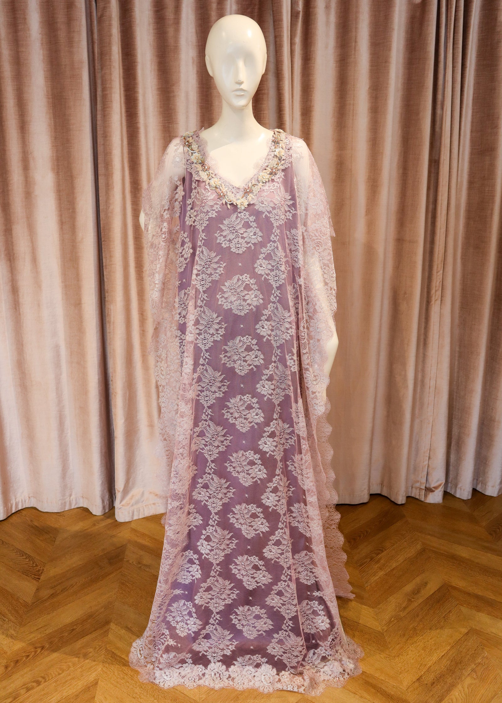 A lavender purple lace caftan dress with floral embroidery and sheer sleeves, displayed on a white mannequin against a pink velvet curtain and wooden parquet floor.