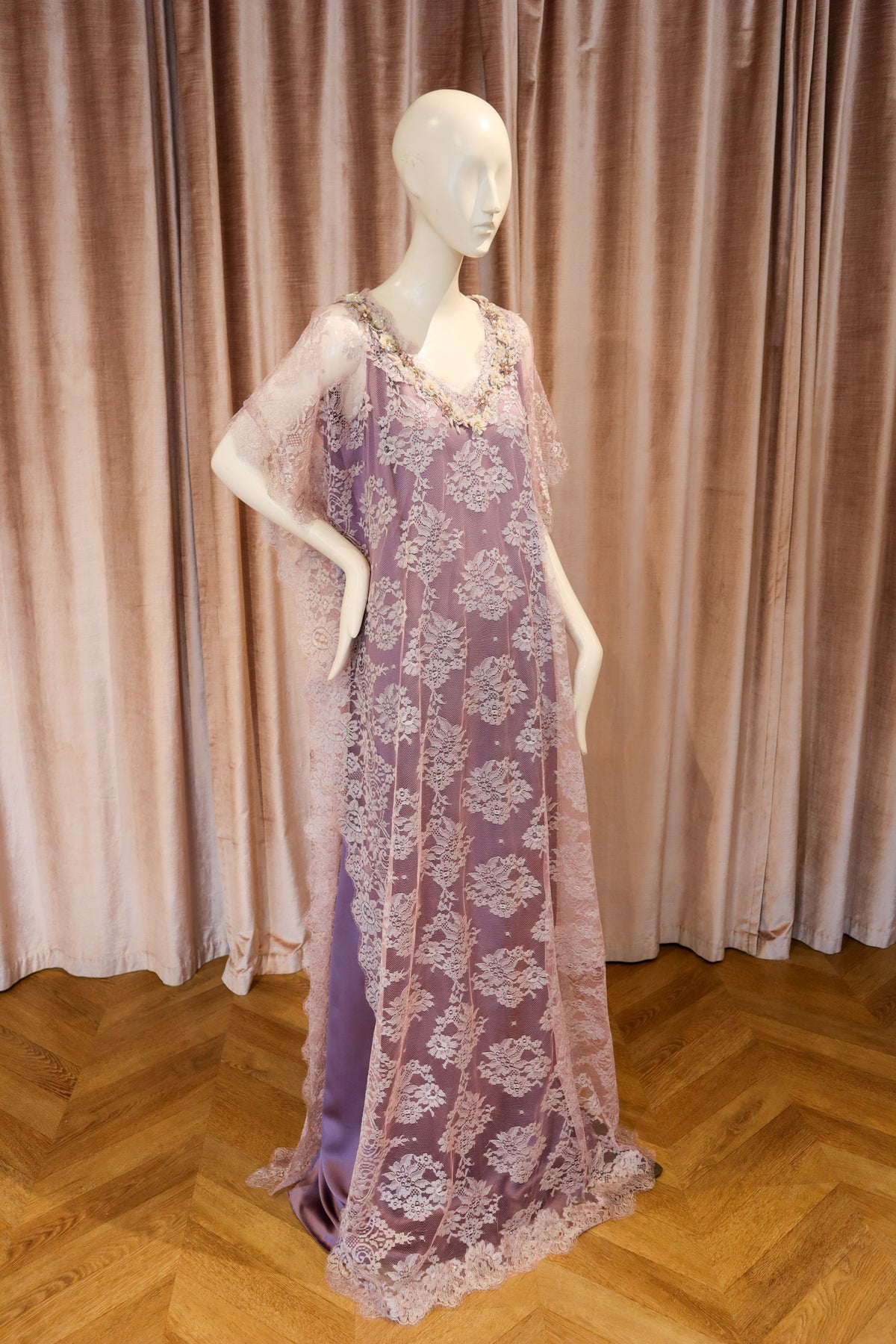 A lavender purple lace caftan dress with floral embroidery and sheer sleeves, displayed on a white mannequin against a pink velvet curtain and wooden parquet floor.