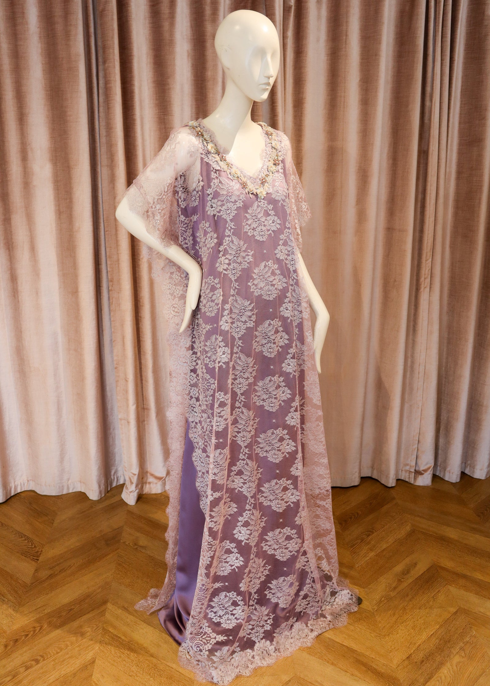 A lavender purple lace caftan dress with floral embroidery and sheer sleeves, displayed on a white mannequin against a pink velvet curtain and wooden parquet floor.