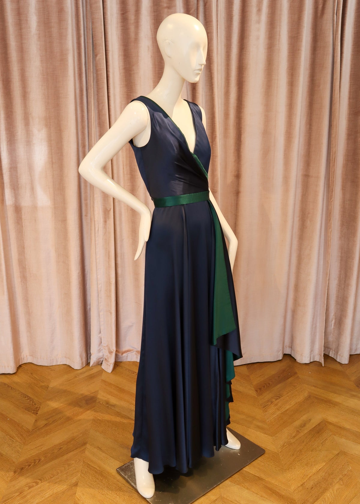 A sleeveless navy blue maxi dress with a deep V-neckline, a green waist sash, and an asymmetrical green side drape, displayed on a white mannequin against a pink curtain backdrop.