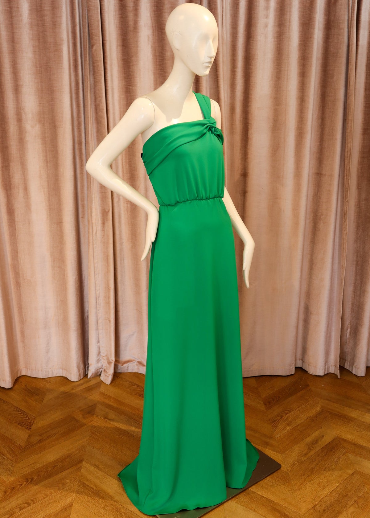 A floor-length emerald green one-shoulder gown displayed on a white mannequin, set against a backdrop of dusty pink curtains and a wooden parquet floor.
