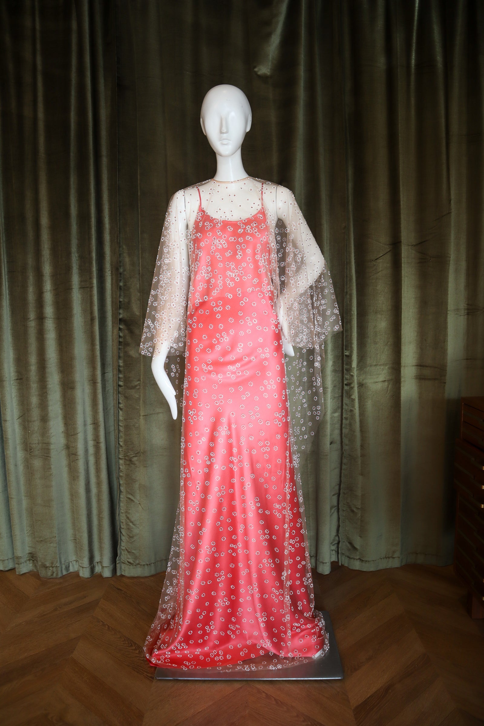 Mannequin wearing a red evening gown with a sheer overlay against a dark curtain background