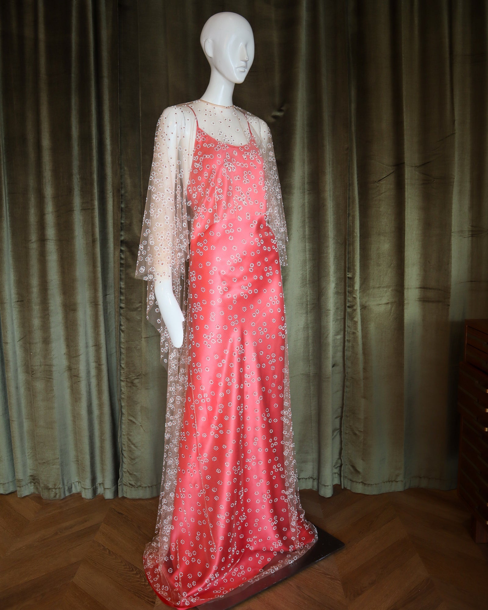 Mannequin wearing a red evening gown with a sheer overlay against a dark curtain background
