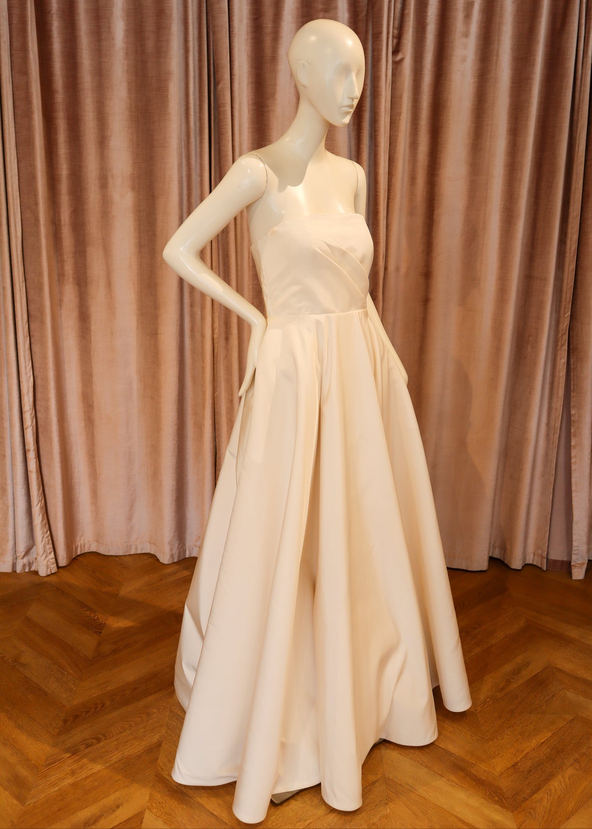 A floor-length off-white evening gown displayed on a white mannequin, set against a backdrop of dusty rose velvet curtains and a wooden parquet floor.