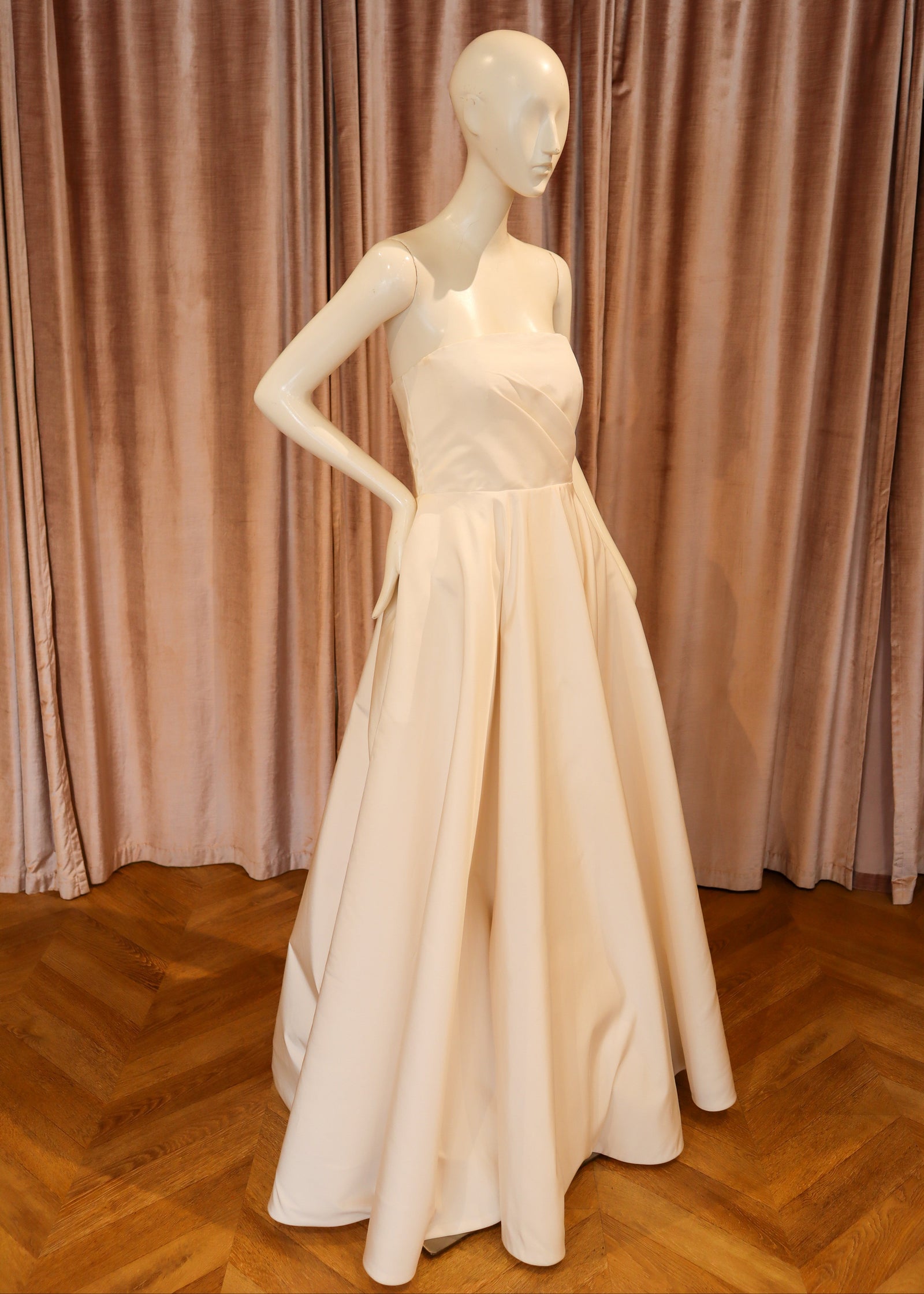 A floor-length off-white evening gown displayed on a white mannequin, set against a backdrop of dusty rose velvet curtains and a wooden parquet floor.