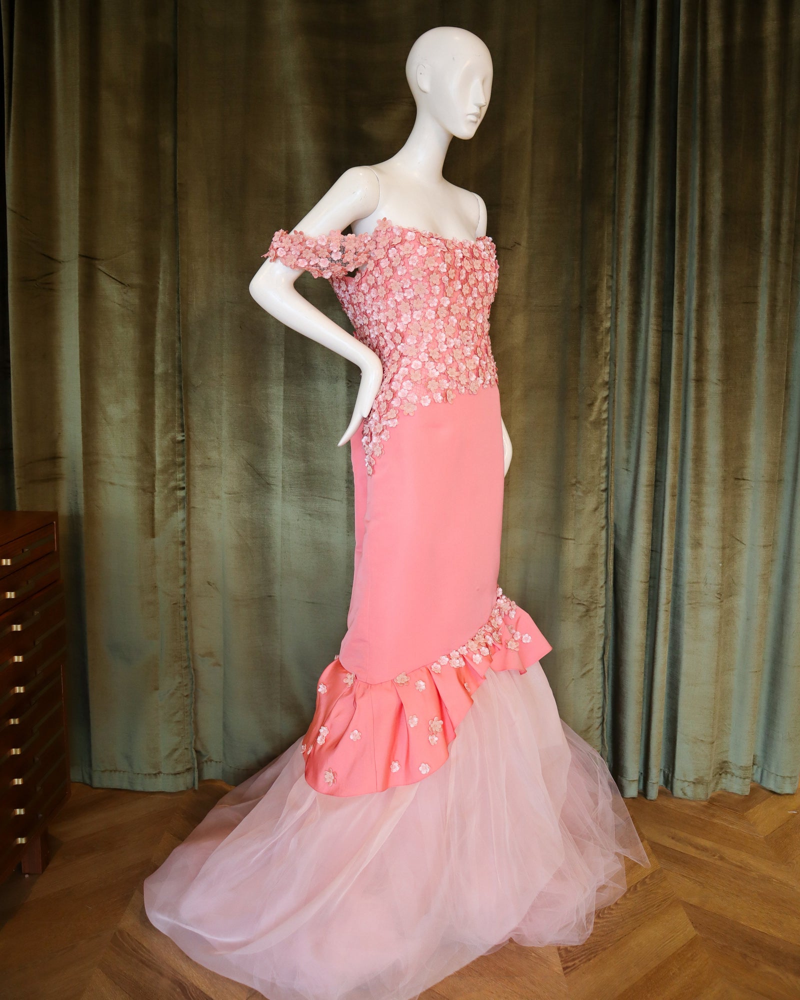 Mannequin wearing a pink evening gown with a textured bodice against a green curtain.