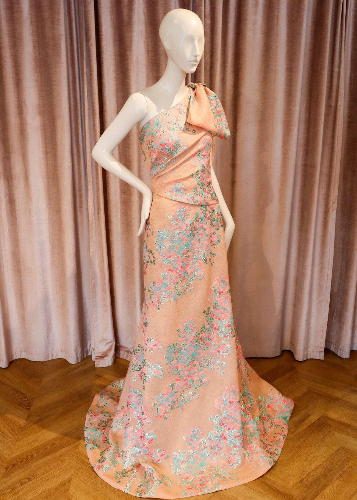 A peach-colored one-shoulder evening gown with a floral print in pink, blue, and green, displayed on a white mannequin against a dusty pink velvet curtain and wooden parquet floor.