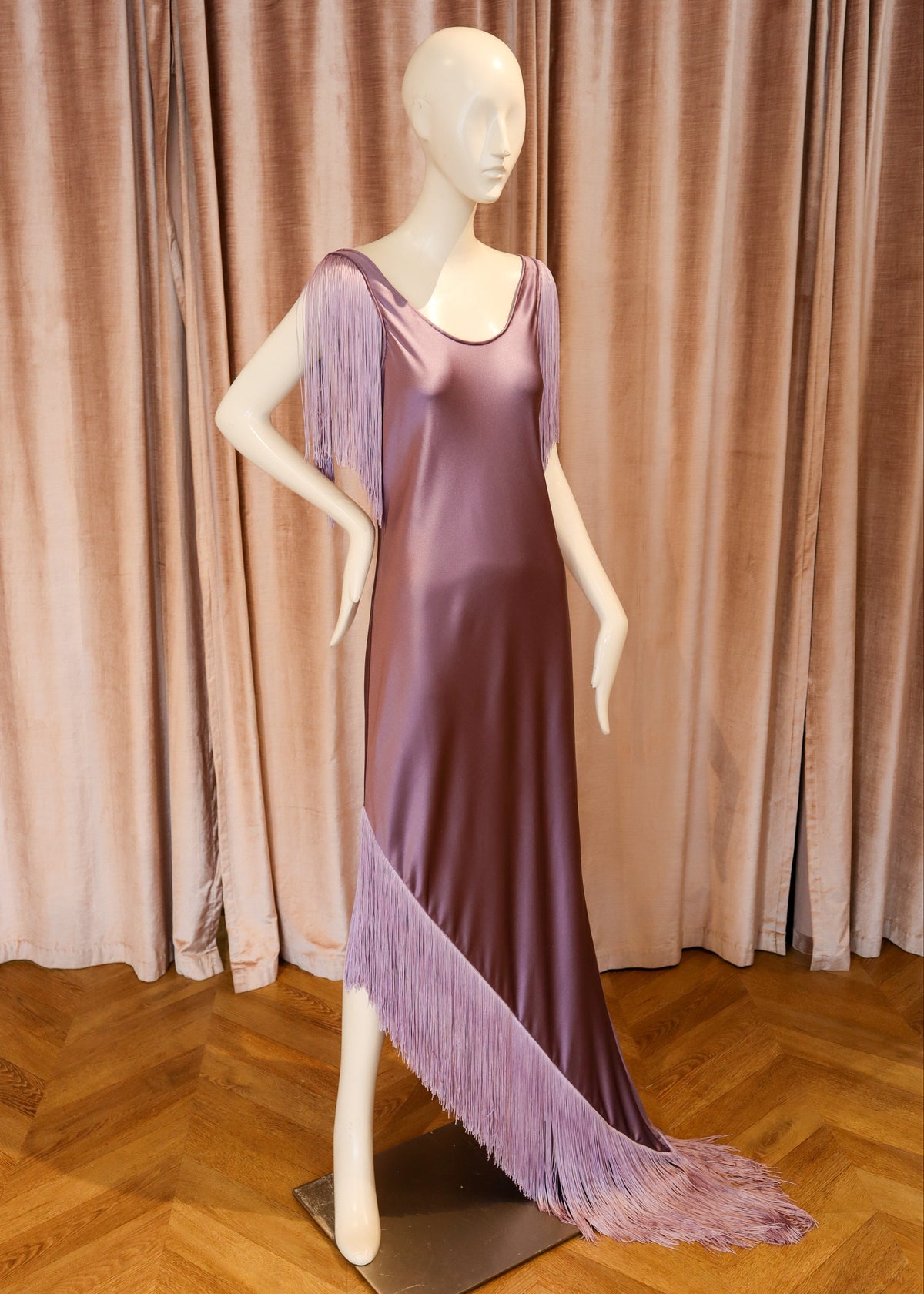 A lavender-purple satin evening gown with fringe detailing, displayed on a female mannequin against a backdrop of dusty pink velvet curtains and a wooden parquet floor.