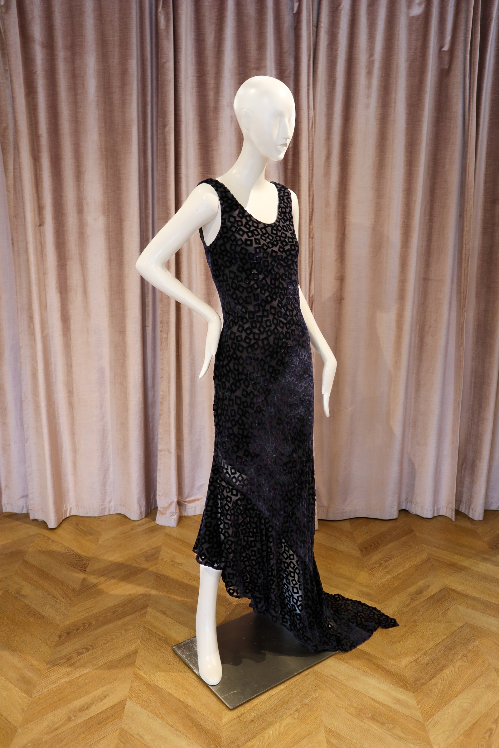 A black beaded evening gown displayed on a white mannequin, set against a backdrop of dusty pink velvet curtains and a light wood herringbone floor.