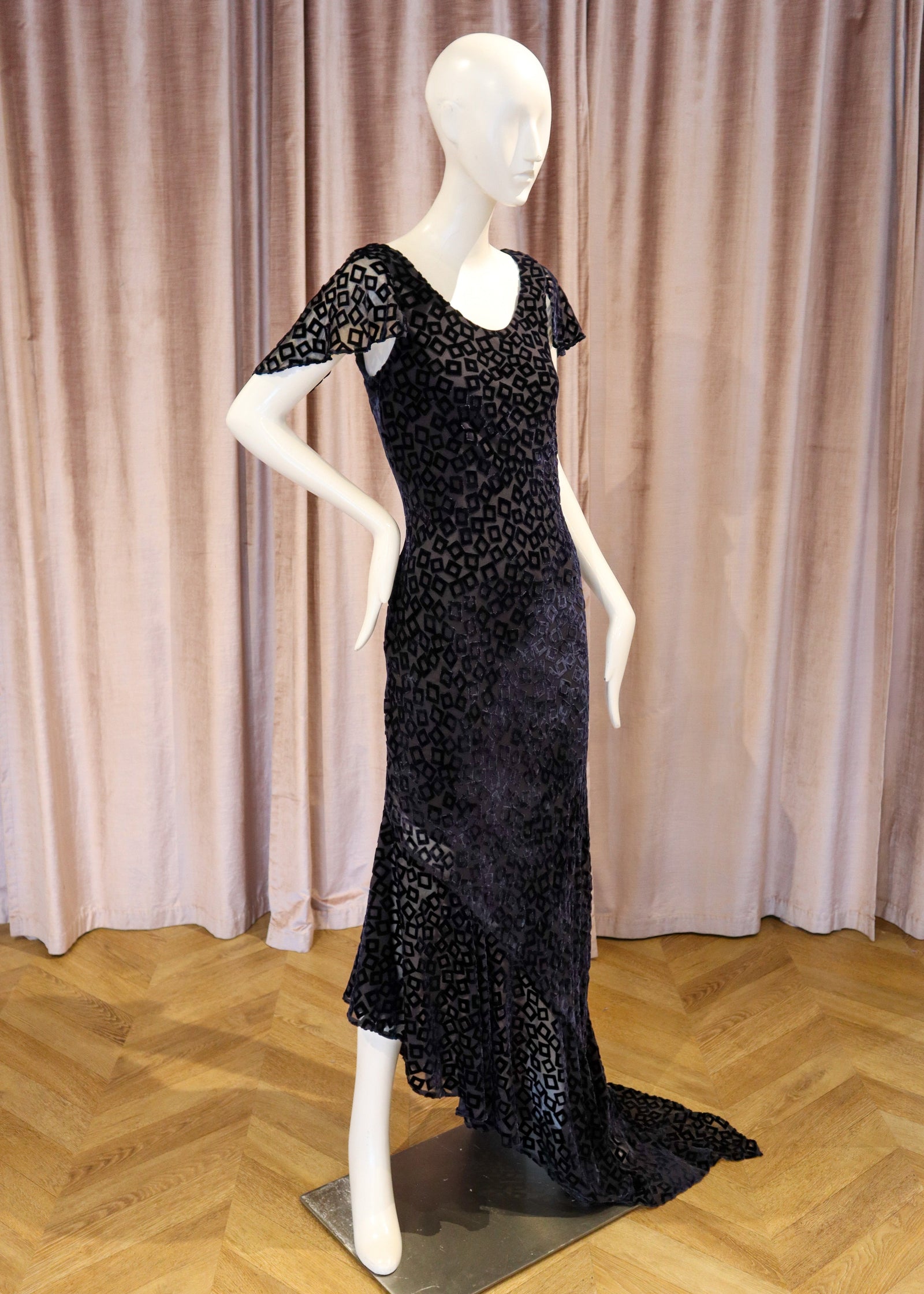 A black beaded evening gown displayed on a white mannequin, set against a backdrop of dusty pink velvet curtains and a light wood herringbone floor.