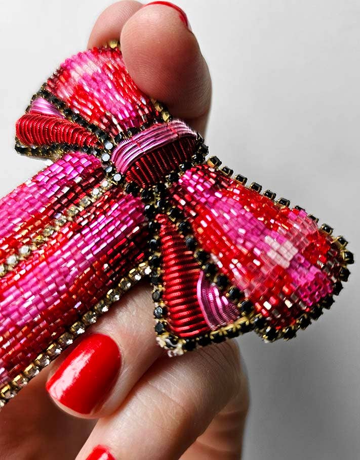 Beaded bow hair brooch held by a hand with red nail polish on a light gray background