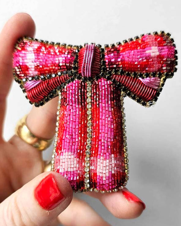 Beaded bow-shaped brooch held by a hand with red nail polish on a light background