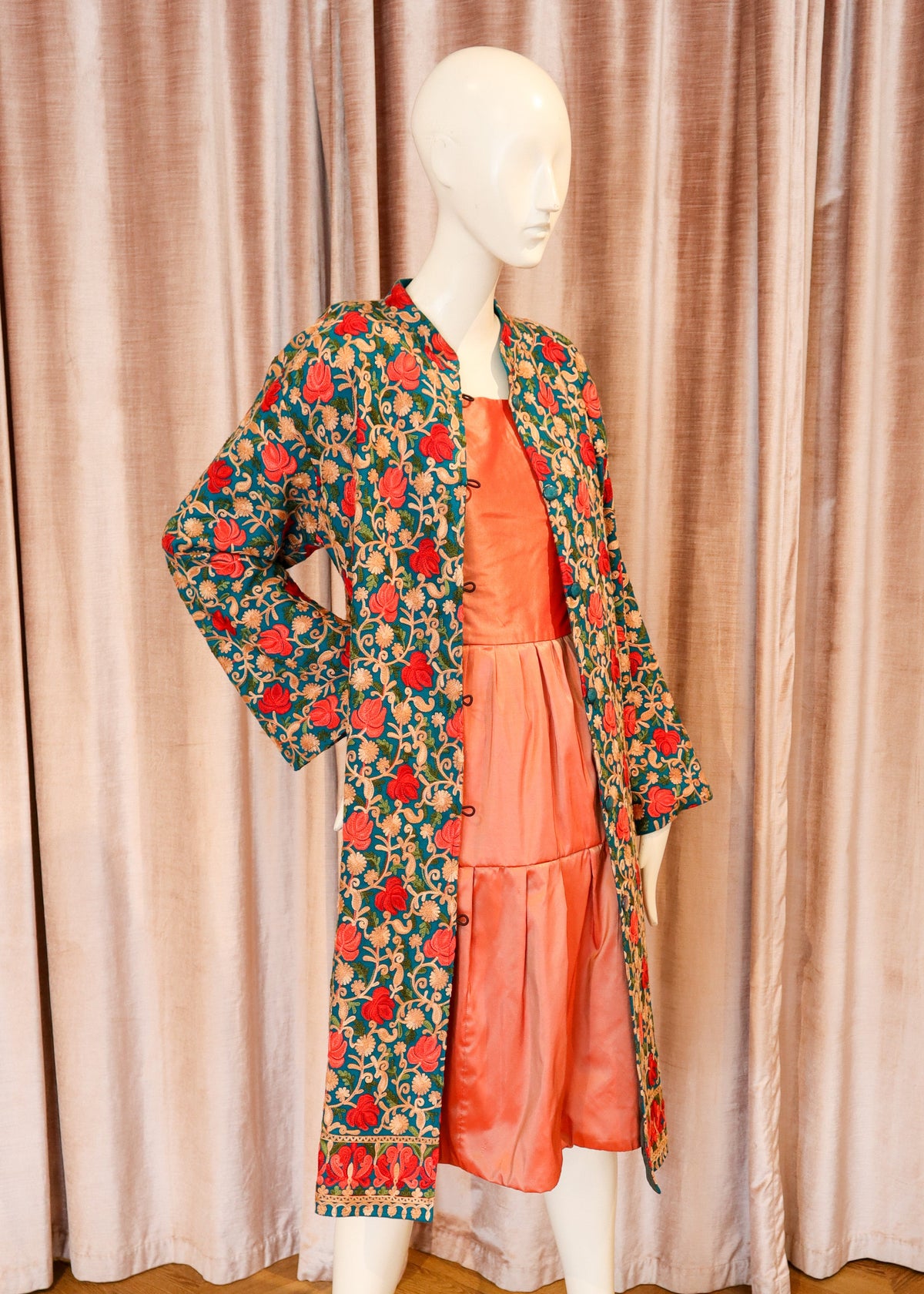 A mannequin wearing a long-sleeved coral pink dress layered under a green floral print jacket, set against a backdrop of pink curtains and a wooden herringbone floor.