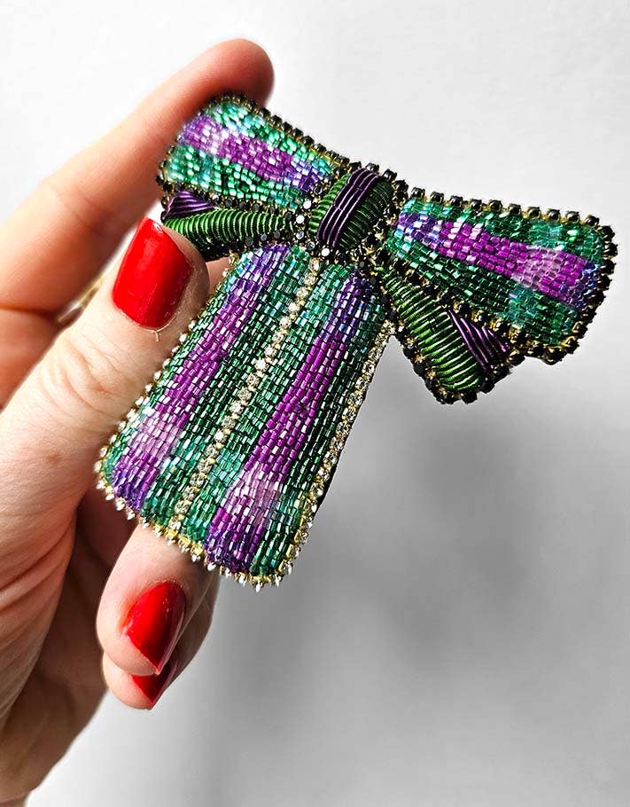Beaded bow brooch held by a hand with red nail polish on a light gray background