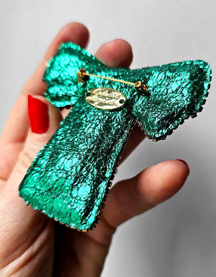 Green metallic butterfly brooch held by a hand with red nail polish on a light gray background