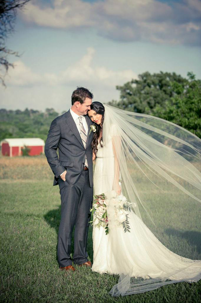Dark haired bride in custom designed short sleeve wedding gown by David Peck, posing with her husband in an open field.