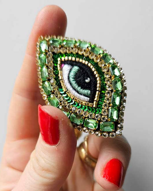 Hand holding a decorative brooch with eye design and green gemstones against a neutral background