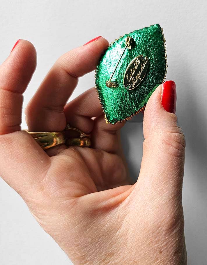 Green eye-shaped brooch held by a hand with red nail polish on a light gray background