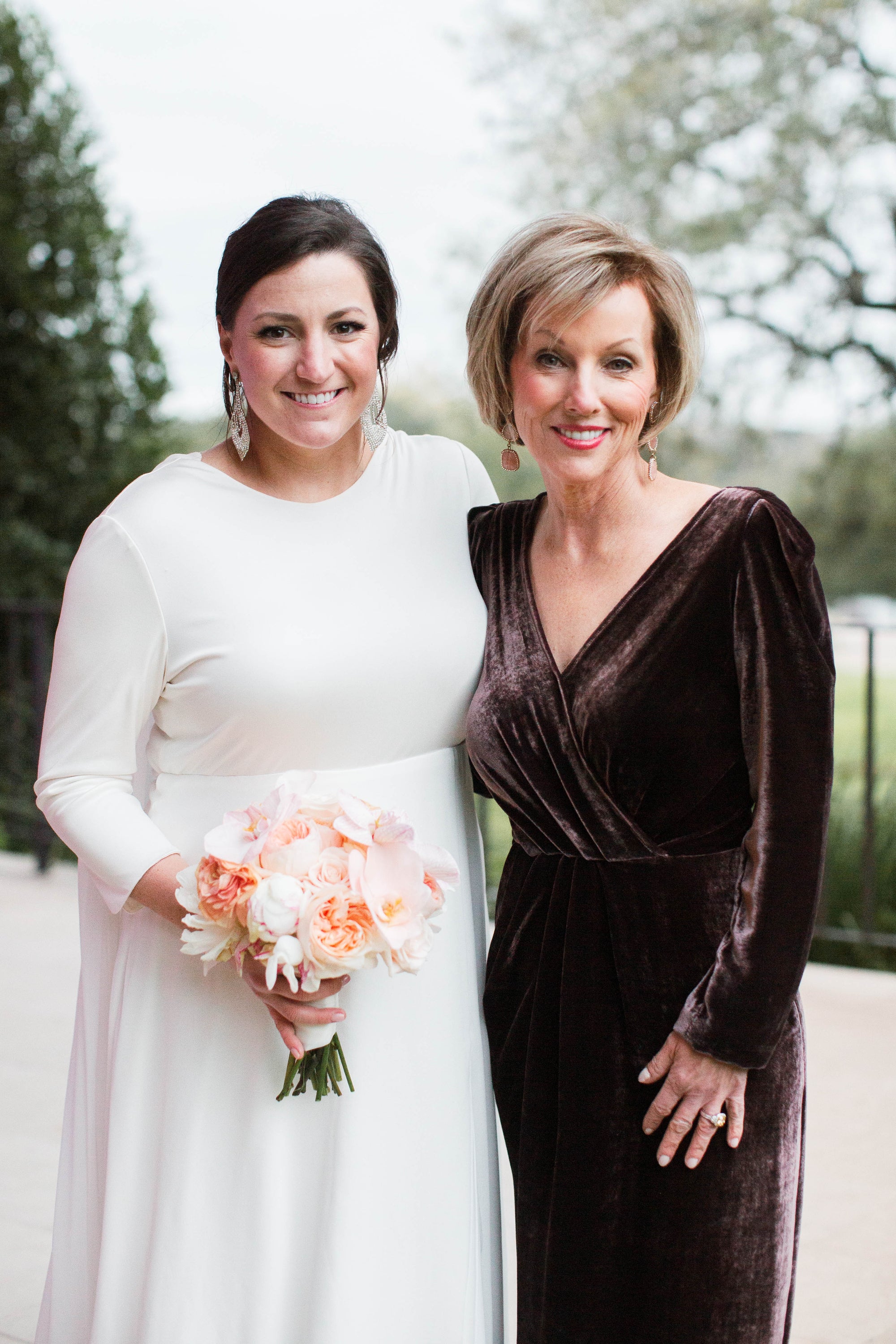 Laura Pipkin in her custom designed long sleeve V-neck and velvet brown mother of the bride dress by David Peck standing next to her daughter Natalie at her wedding in Texas. 